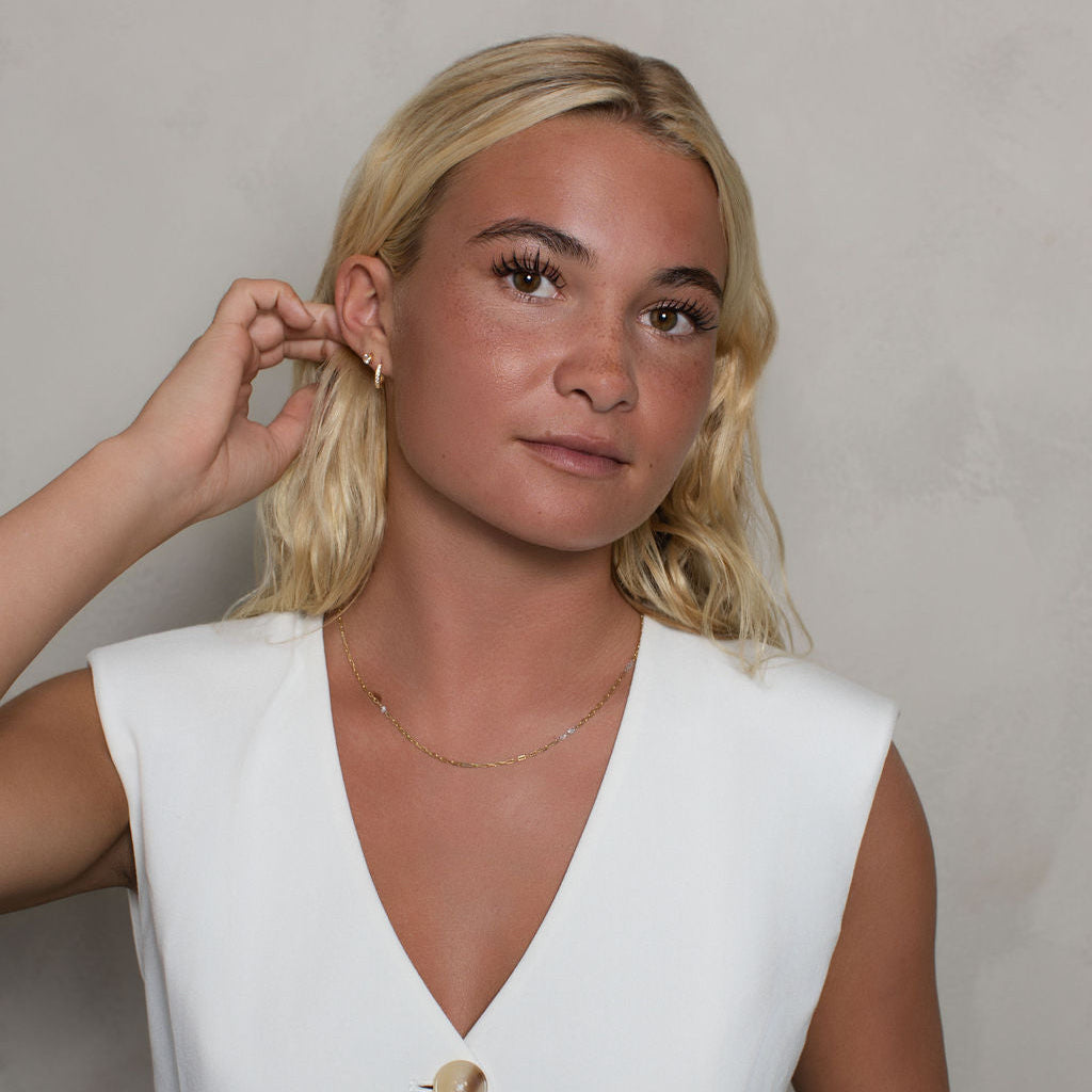 A blonde woman wearing a white sleeveless V-neck top with gold buttons, posing against a neutral light gray wall.