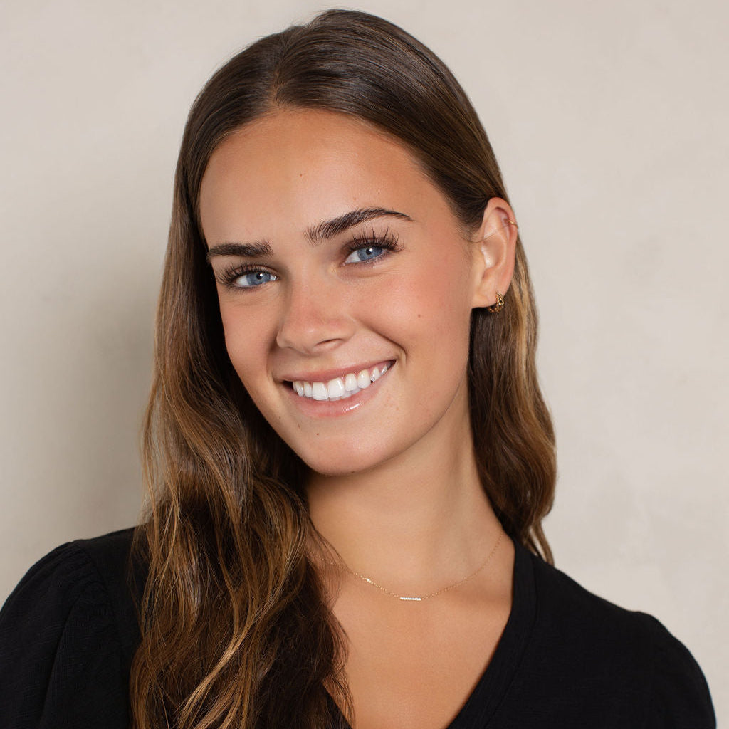 A smiling young woman with long wavy brown hair wearing a black V-neck top with scalloped short sleeves, photographed against a plain light beige wall.
