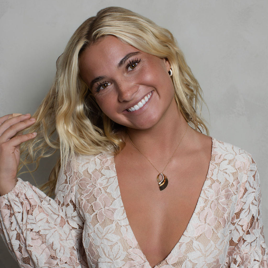 A smiling blonde woman wearing a light beige and white floral lace long-sleeve wrap dress, accessorized with a delicate gold crescent moon pendant necklace, posing against a neutral gray wall.