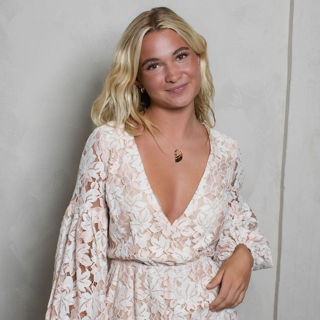 A woman with blonde hair wearing a light pink and white floral lace dress with a deep V-neck and long puff sleeves, standing against a textured light gray wall.