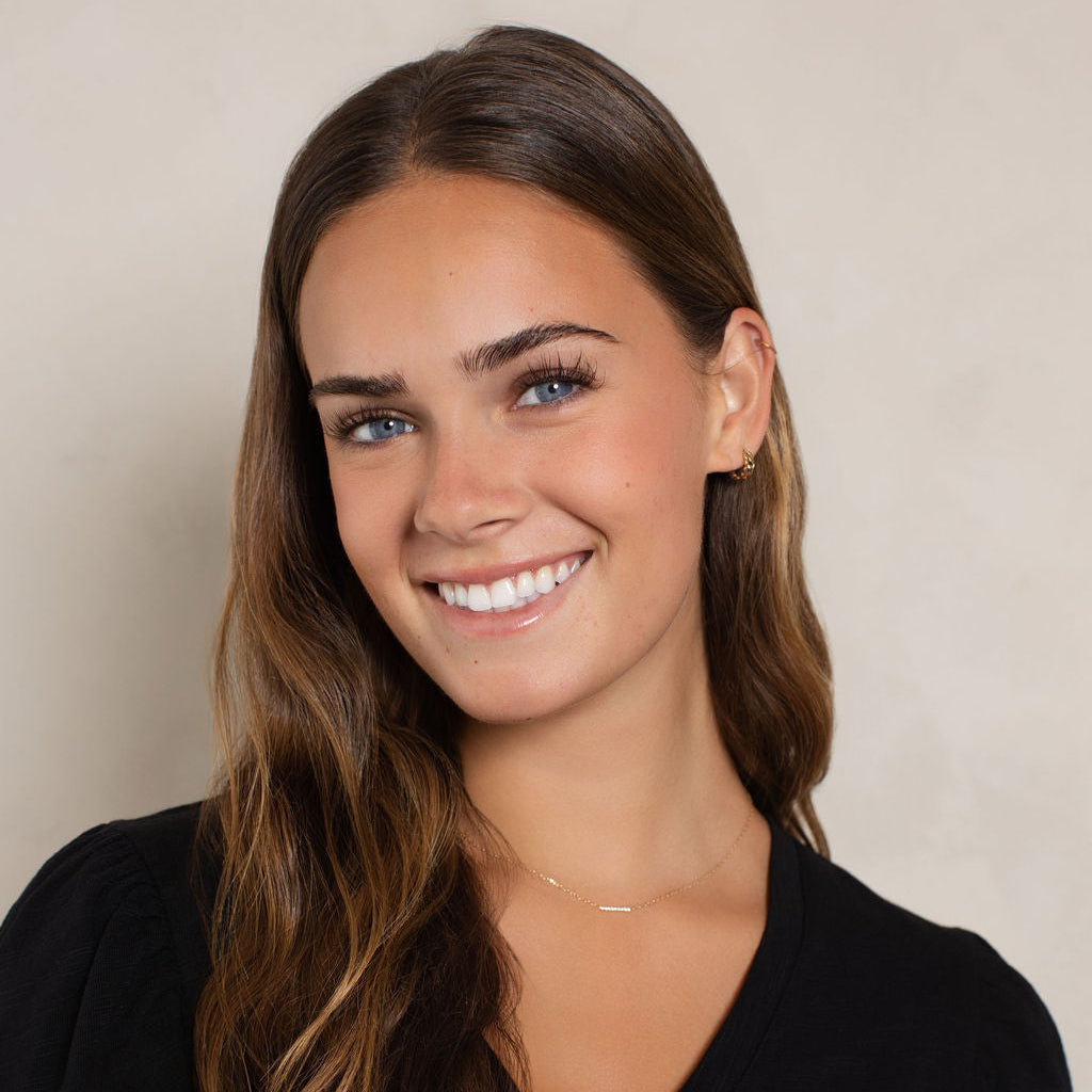 A smiling young woman with long wavy brown hair wearing a black V-neck top with scalloped short sleeves, photographed against a plain light beige wall.