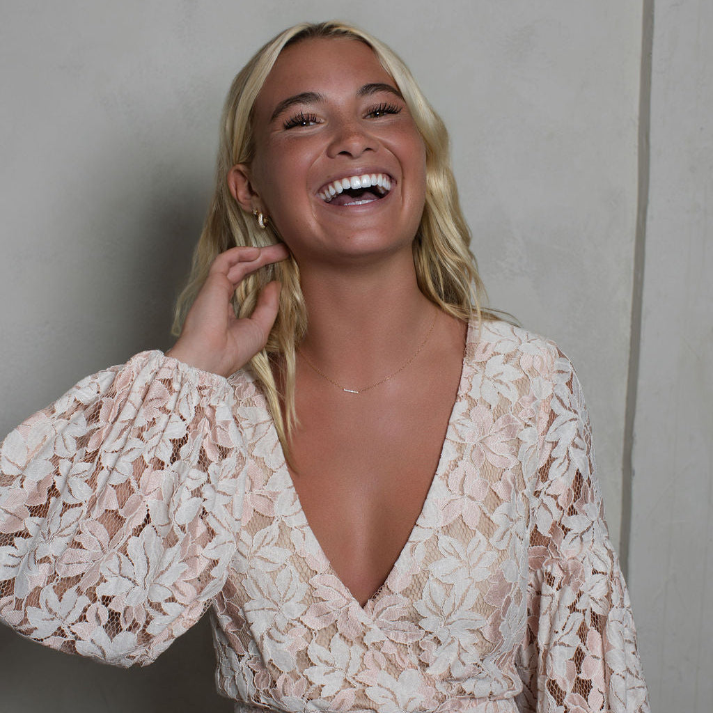 A smiling blonde woman wearing a light beige and white floral lace long-sleeve dress, posing against a textured gray wall.