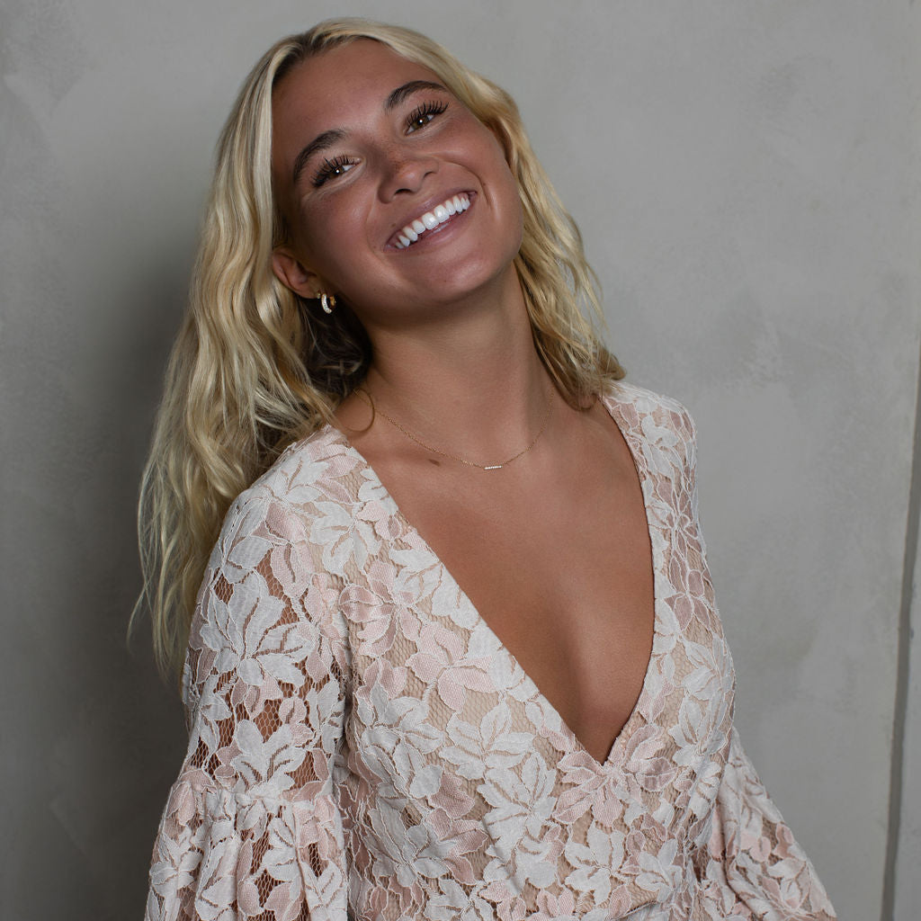 A smiling blonde woman wearing a light beige and white floral lace long-sleeve dress with a deep V-neck, posing against a textured gray wall.
