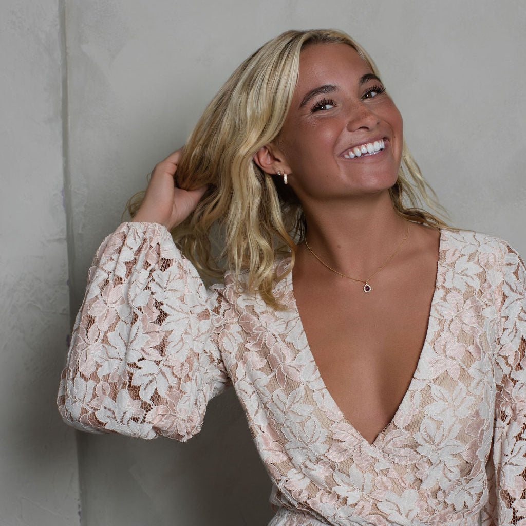A smiling blonde woman wearing a light beige and white floral lace dress with a deep V-neck and long puff sleeves, posing against a textured gray wall.