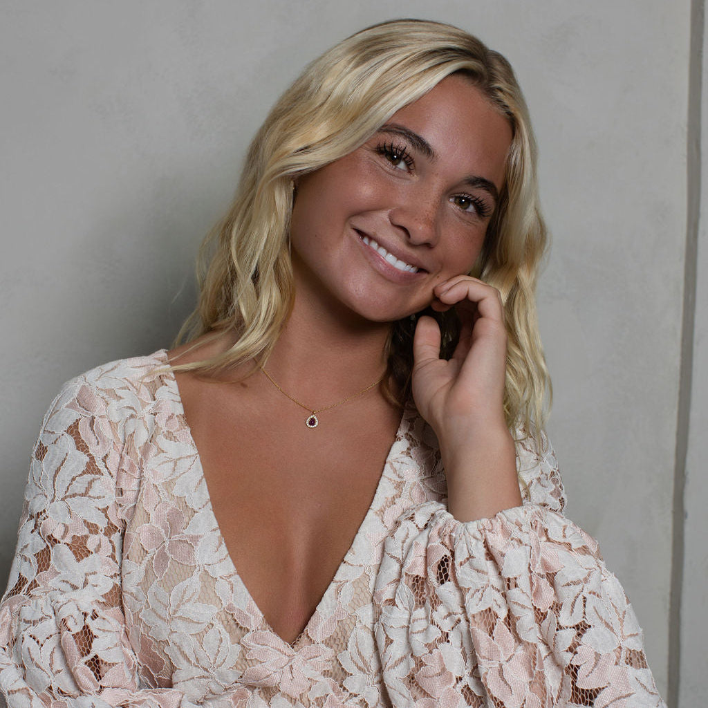 A smiling blonde woman wearing a light pink and white floral lace long-sleeve top, posing against a neutral light gray wall.