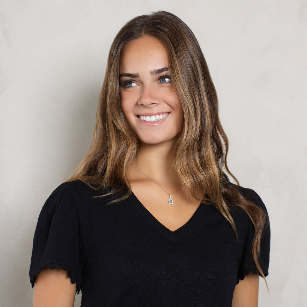 A smiling woman with long brown hair wearing a black short-sleeve V-neck top, standing against a light gray textured wall.
