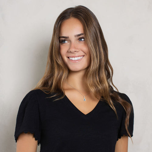 A smiling woman with long brown hair wearing a black short-sleeve V-neck top, standing against a light gray textured wall.