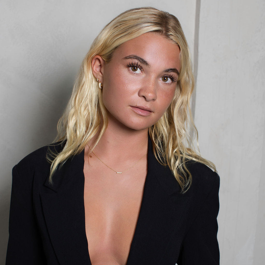 A blonde woman wearing a black open-front blazer with a very deep V-neckline, posed against a light gray textured wall.
