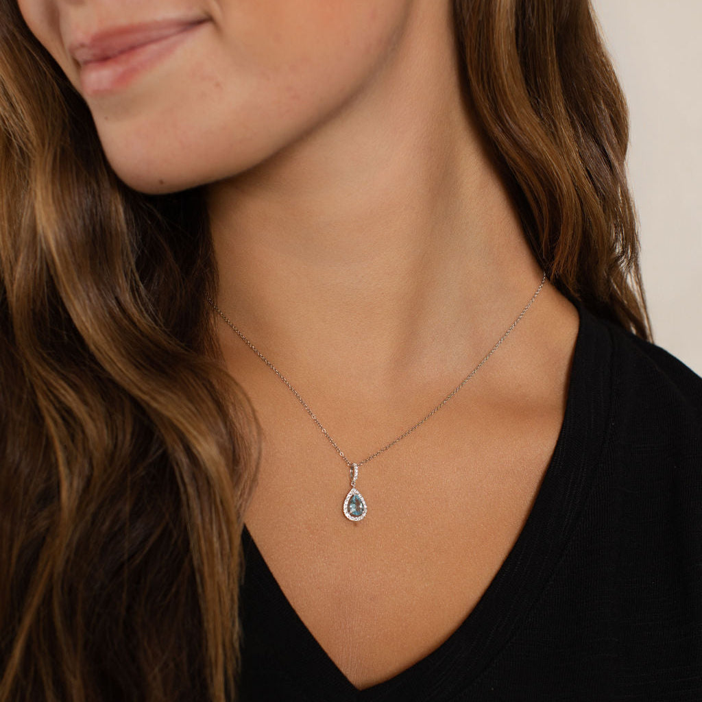 A close-up of a woman wearing a delicate silver pendant necklace with a teardrop-shaped clear gemstone, paired with a black V-neck top.