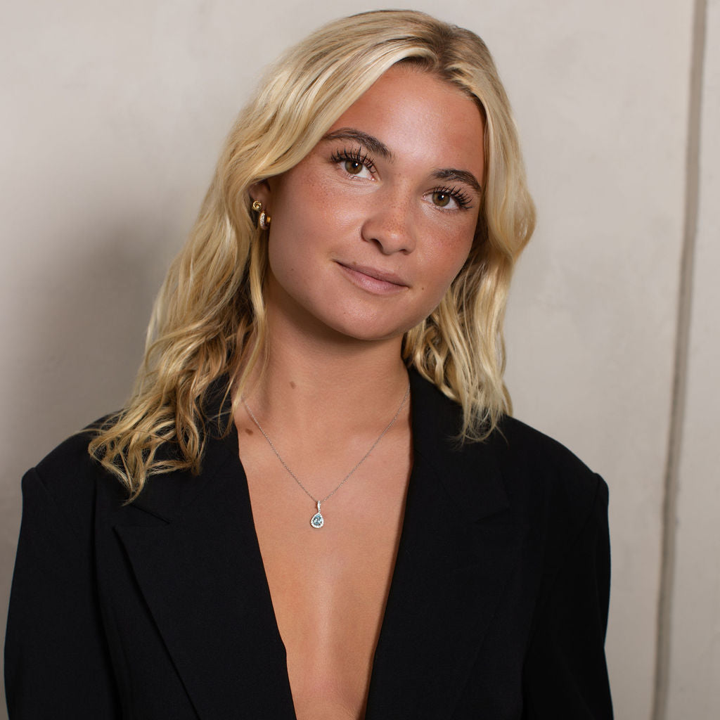 A blonde woman wearing a black blazer and a delicate silver pendant necklace, photographed against a neutral beige wall.