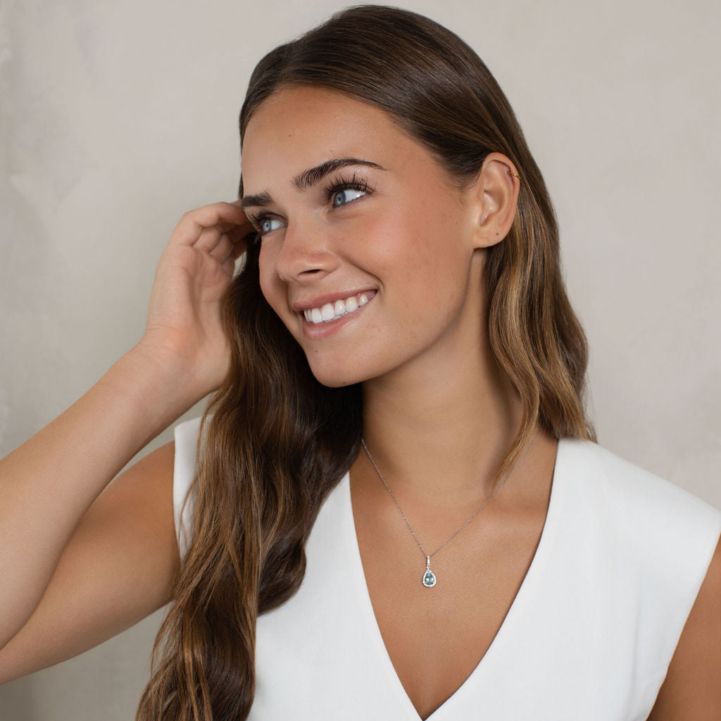 A smiling woman with long wavy brown hair wears a delicate silver necklace with a small light blue stone pendant, paired with a white sleeveless top. She is posing against a soft, textured beige background.