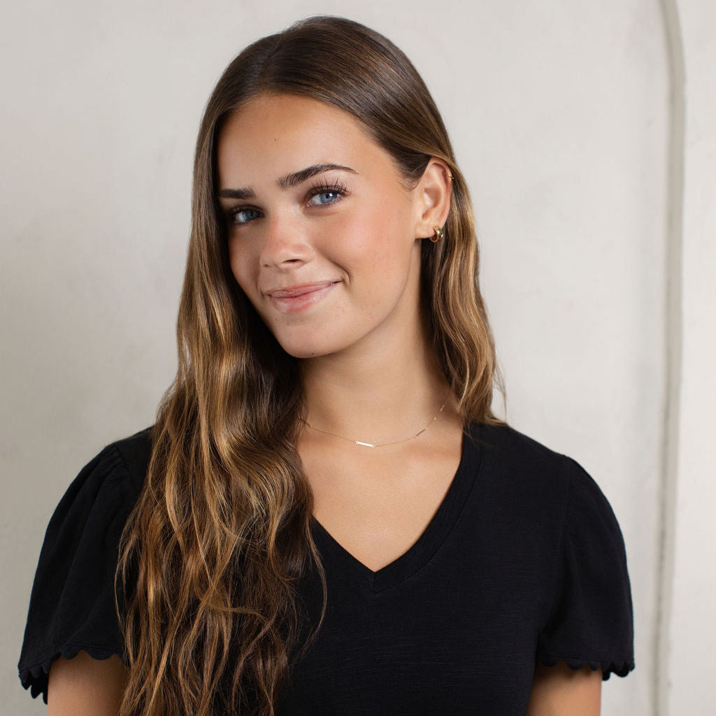 A woman with long, wavy brown hair wearing a black short-sleeve V-neck t-shirt with scalloped edges, standing against a light gray textured wall.