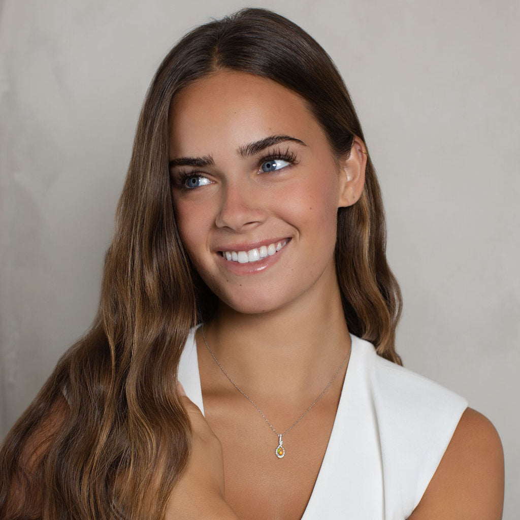 A smiling woman with long wavy brown hair wearing a white sleeveless top, a delicate gold necklace, and stacked silver bracelets, posed against a neutral light gray textured background.