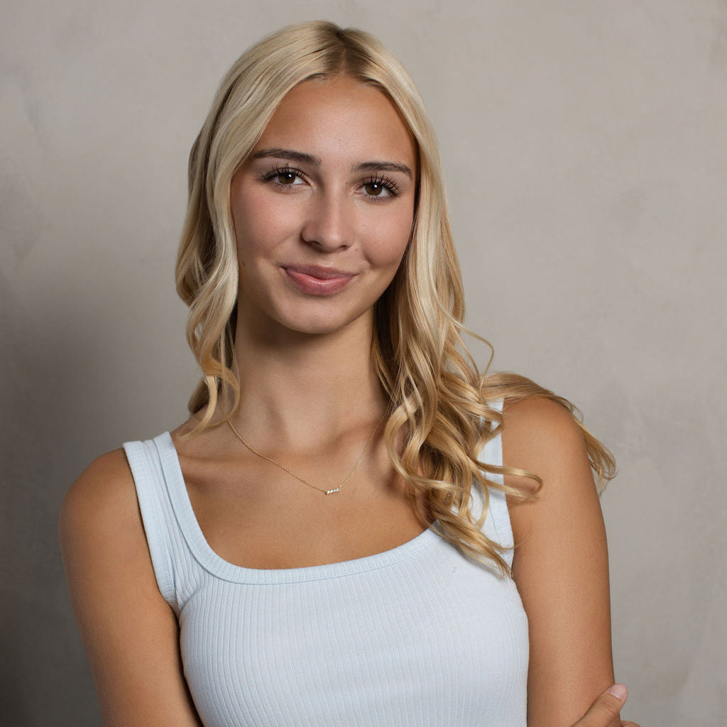 A young blonde woman with wavy hair wearing a light blue ribbed tank top, posing with her arms crossed against a neutral textured gray background.