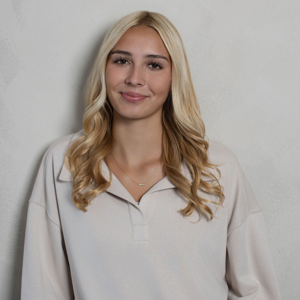 A blonde woman wearing a beige long-sleeve polo sweatshirt with white stripes at the hem, standing against a light gray textured wall.
