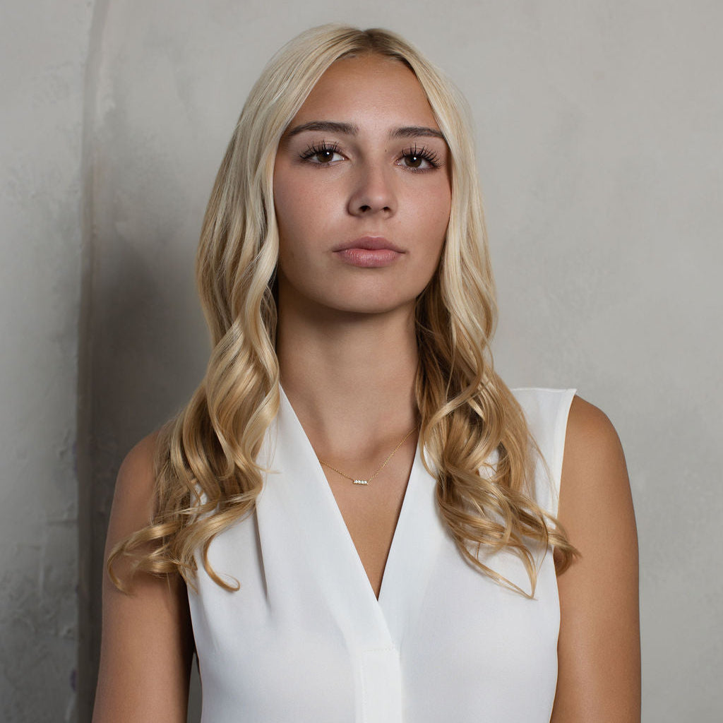 A blonde woman with wavy hair wearing a white sleeveless V-neck top, standing against a textured gray wall.