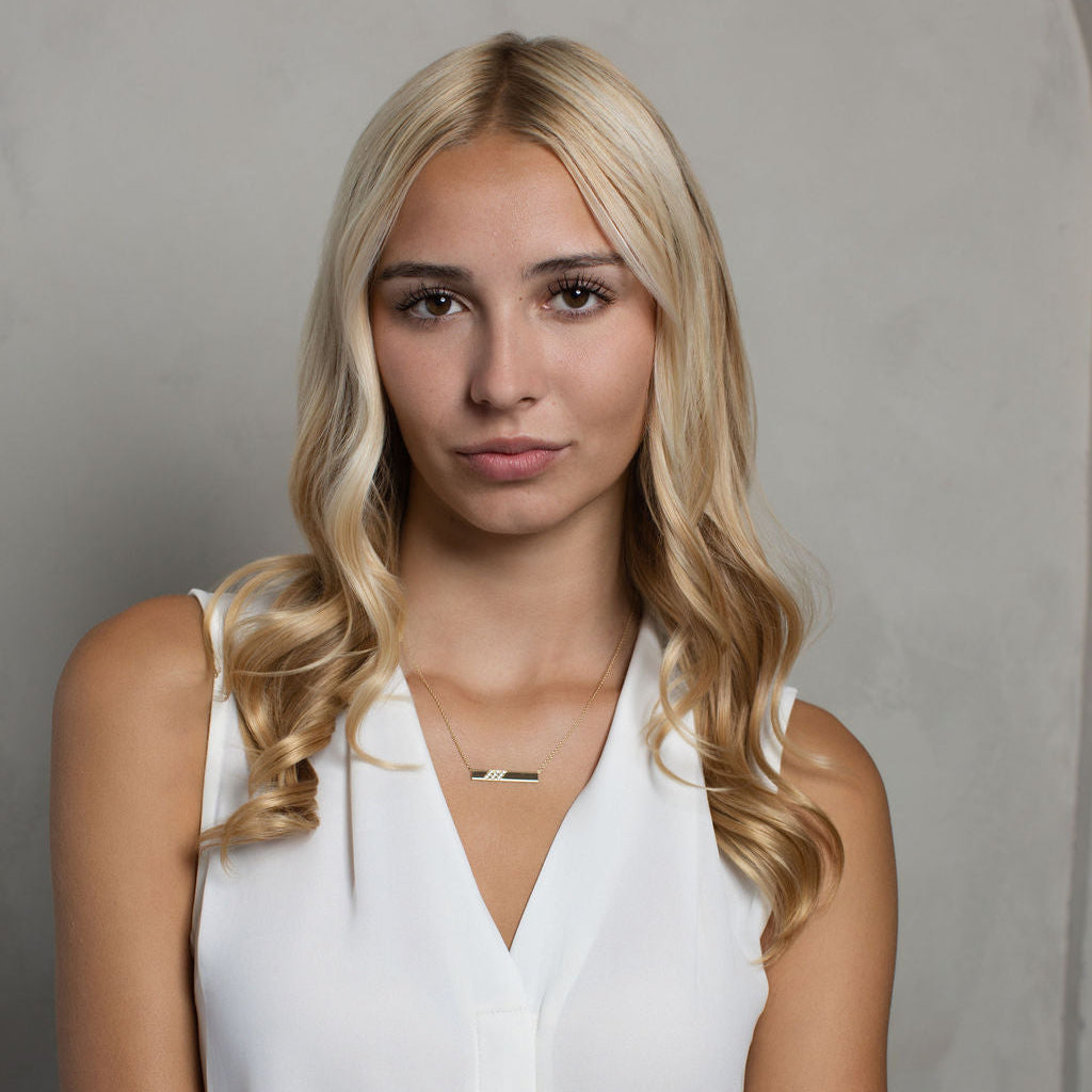 A blonde woman wearing a white sleeveless top and a delicate gold bar necklace, posing against a neutral gray studio background.