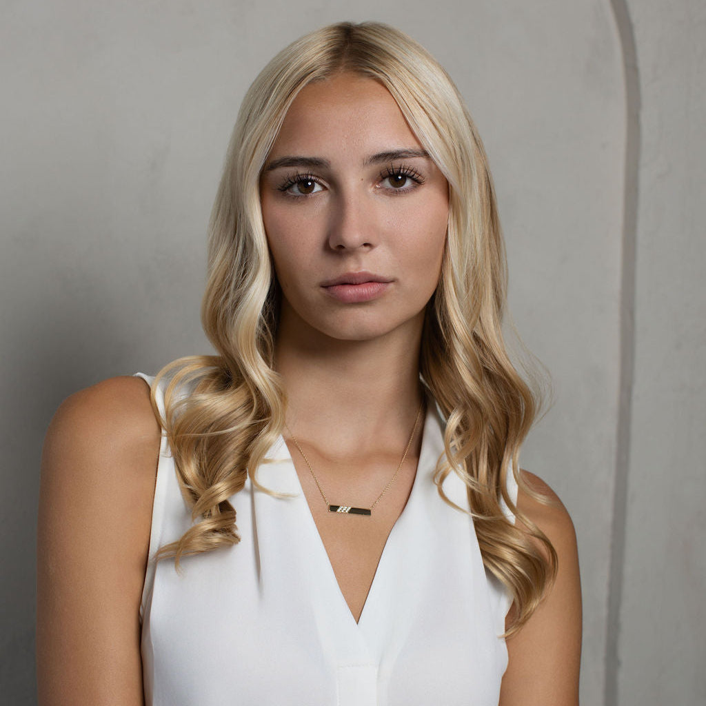 A blonde woman with wavy hair wearing a white sleeveless V-neck top and a delicate gold necklace, posing against a textured gray wall.