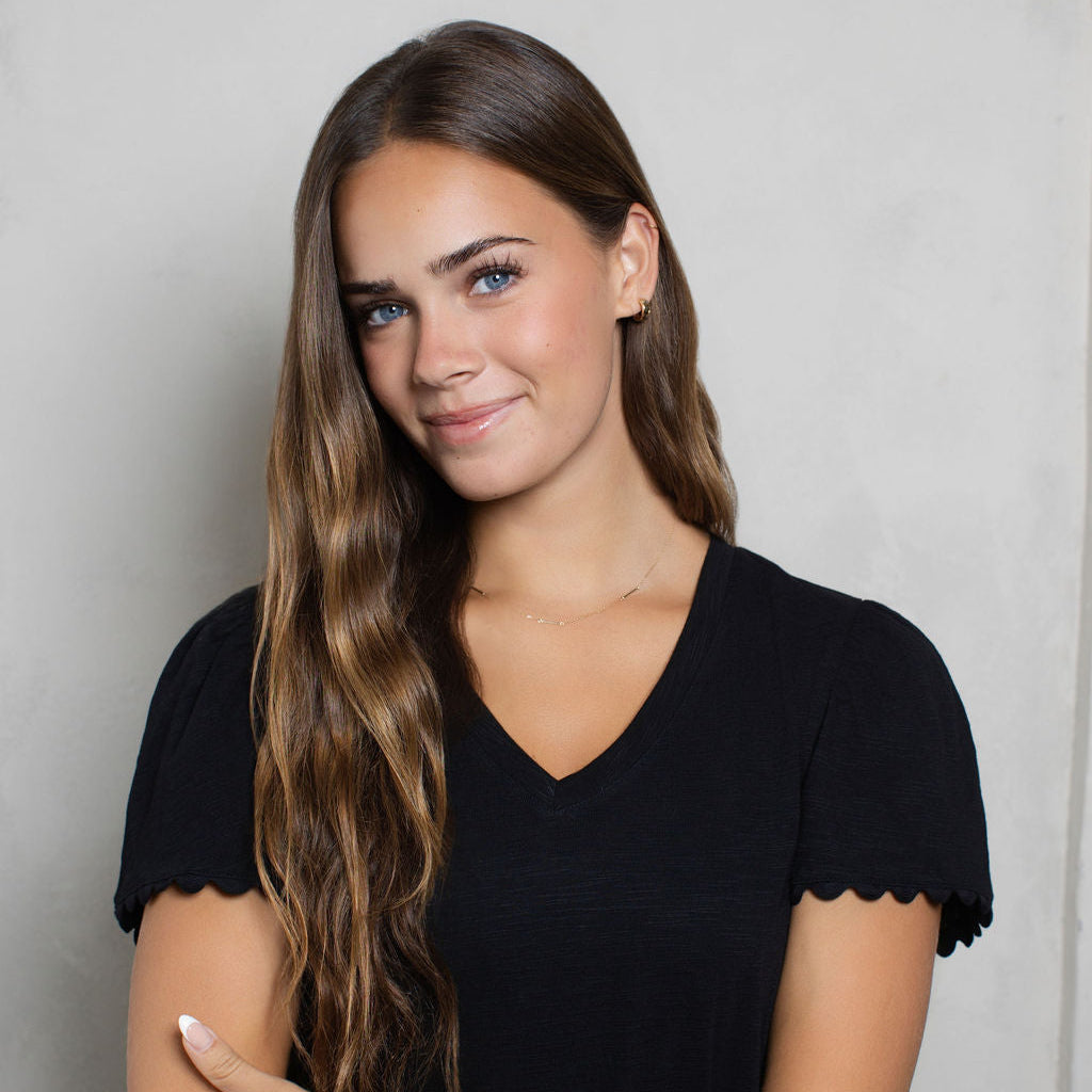 A woman with long, wavy brown hair wearing a black short-sleeve V-neck top with scalloped edges, posing with her arms crossed against a light gray textured background.