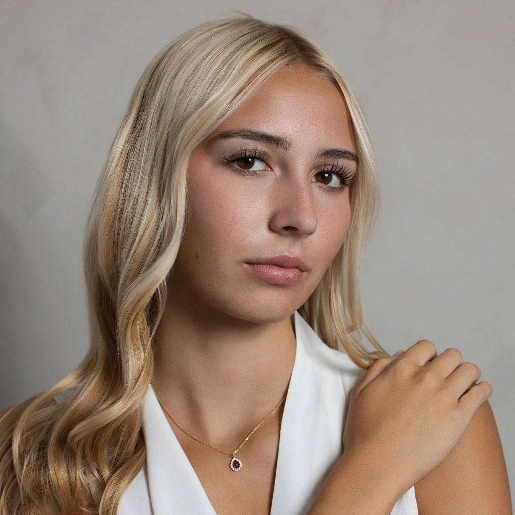 A blonde woman wearing a delicate gold necklace with a small dark circular pendant and a thin red beaded bracelet, posing in a white sleeveless top against a neutral gray background.