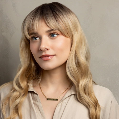 A woman with long blonde wavy hair and straight bangs, wearing a beige button-up shirt and a gold bar necklace, photographed against a textured gray studio background.