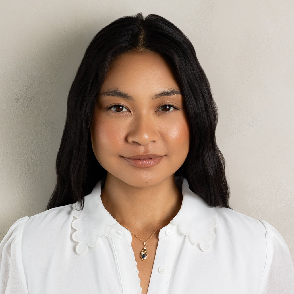 A woman with long black hair wearing a white button-up blouse with a scalloped Peter Pan collar, photographed against a plain light beige wall.