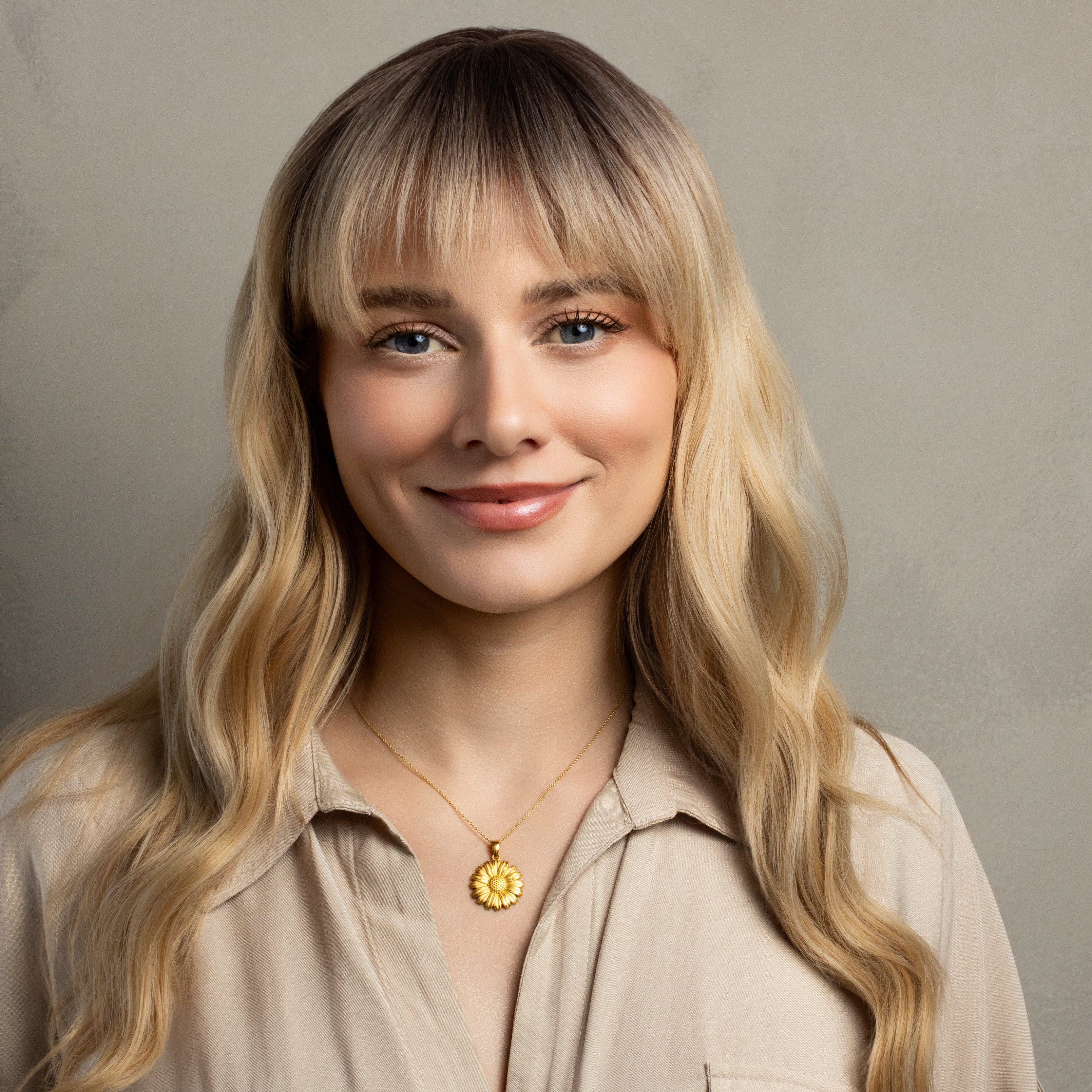A woman with long blonde wavy hair and straight bangs wearing a beige button-up shirt and a gold circular pendant necklace, photographed against a neutral textured gray background.