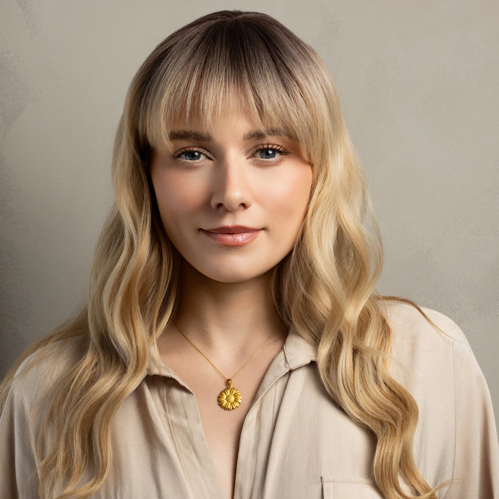 A woman with long wavy blonde hair and dark roots wearing a beige button-up shirt and a gold sun pendant necklace, set against a neutral textured gray background.
