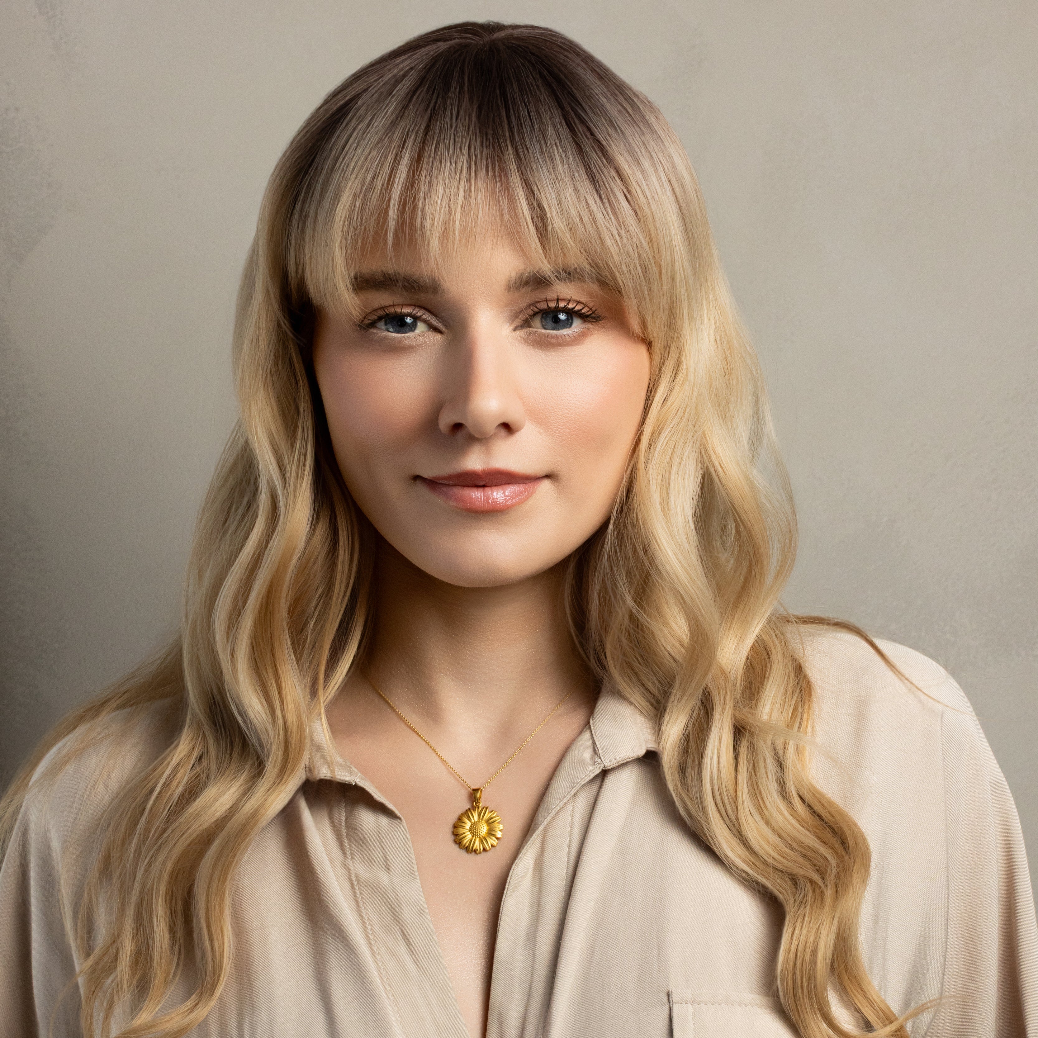A woman with long wavy blonde hair and dark roots wearing a beige button-up shirt and a gold sun pendant necklace, set against a neutral textured gray background.