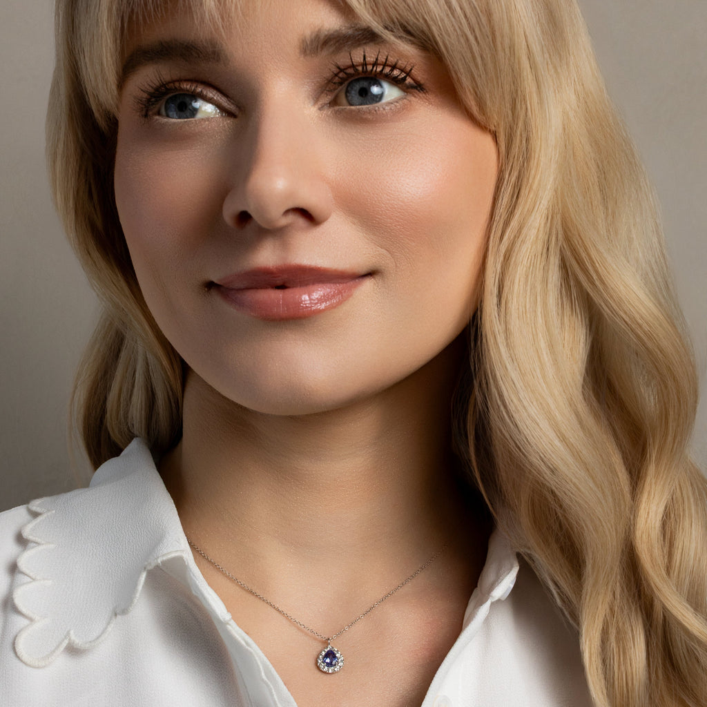 A close-up portrait of a woman with blonde hair wearing a delicate silver necklace with a small blue gemstone pendant, paired with a white collared blouse.
