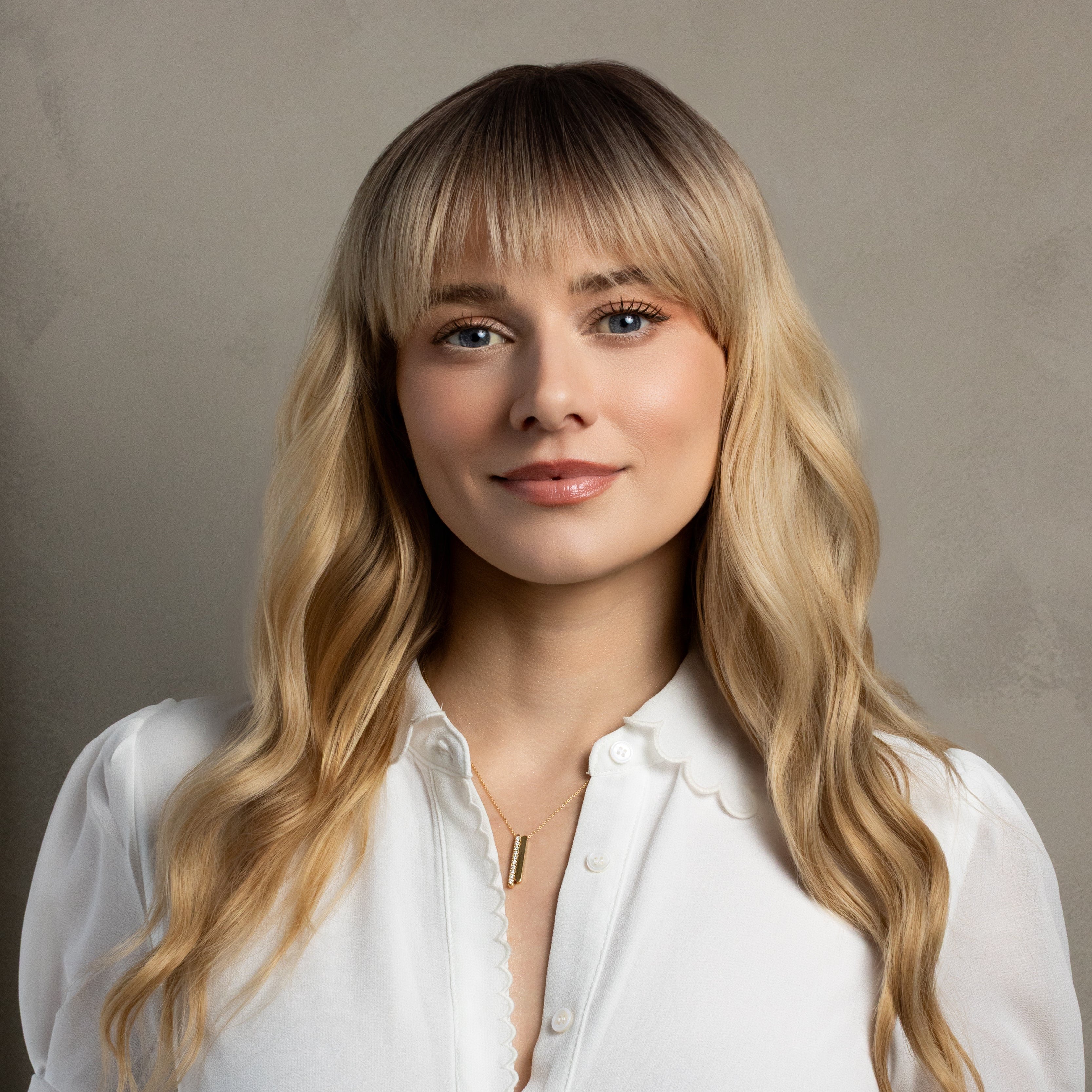 A woman with long blonde wavy hair and straight bangs wearing a white short-sleeve blouse with scalloped edges, photographed against a neutral textured beige background.