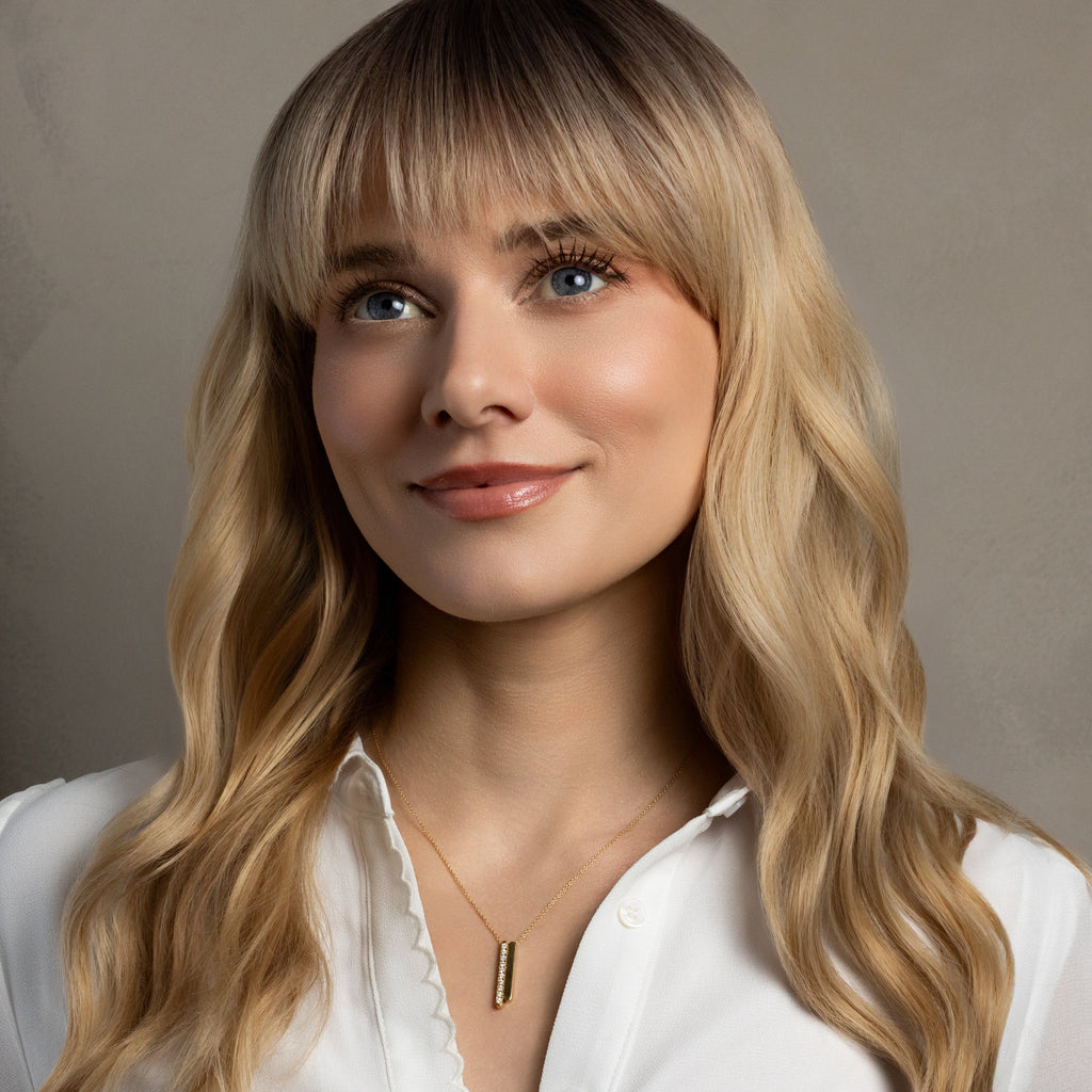A close-up portrait of a woman with long blonde wavy hair and bangs, wearing a white collared blouse and a delicate gold necklace with a vertical bar pendant, set against a neutral gray background.