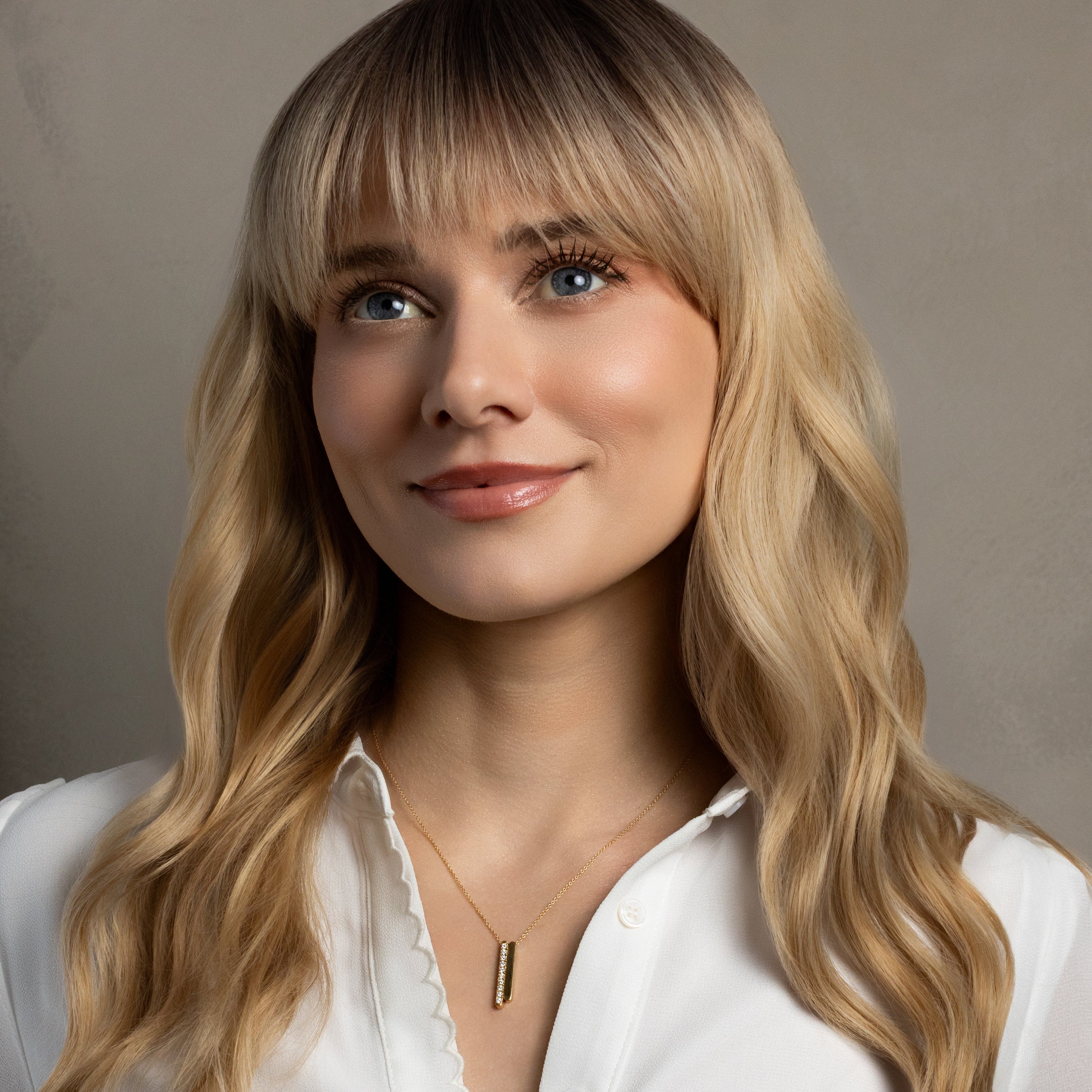 A close-up portrait of a woman with long blonde wavy hair and bangs, wearing a white collared blouse and a delicate gold necklace with a vertical bar pendant, set against a neutral gray background.