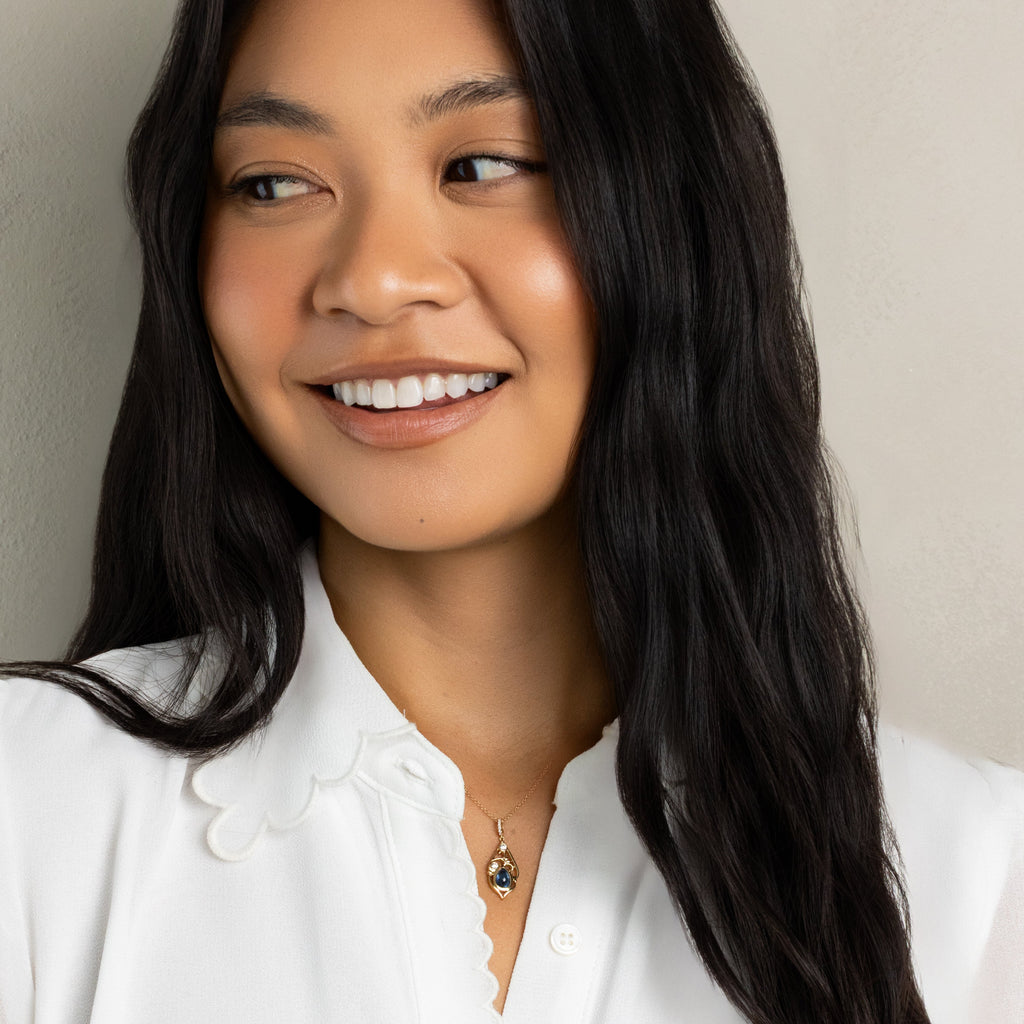 A smiling woman with long dark hair wearing a white button-up blouse and a delicate gold pendant necklace, photographed against a plain light beige wall.