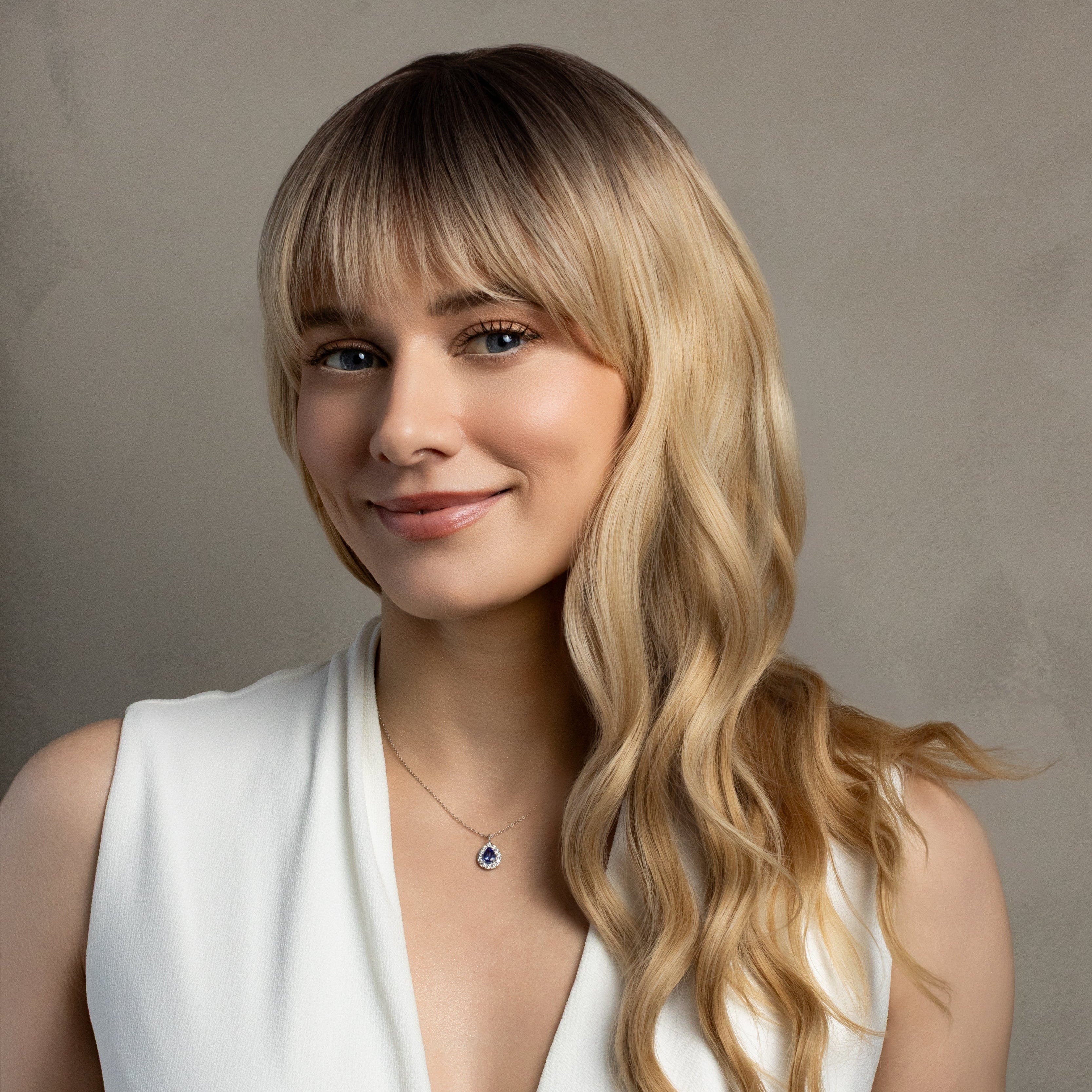 A woman with blonde wavy hair and bangs wearing a white sleeveless V-neck top, photographed against a neutral textured gray background.