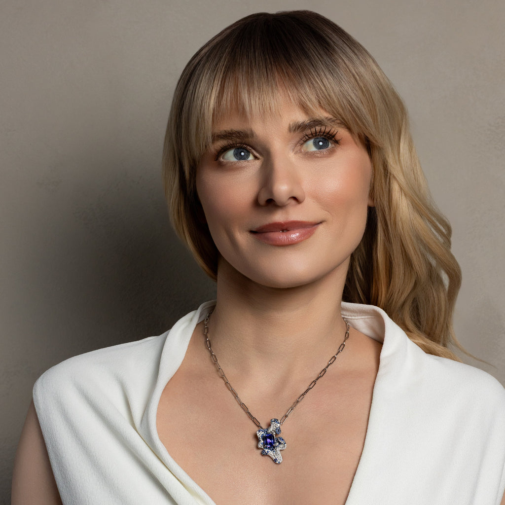 A woman with blonde hair and blue eyes wearing a silver-toned necklace with a blue and white starburst pendant, dressed in a white top against a neutral gray background.
