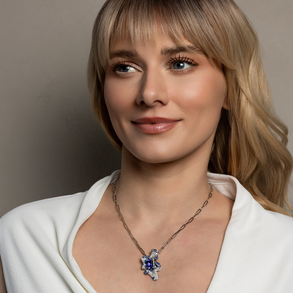 A close-up portrait of a woman with blonde hair wearing a silver-toned chain necklace with a star-shaped pendant featuring a deep blue center stone and small clear stones.