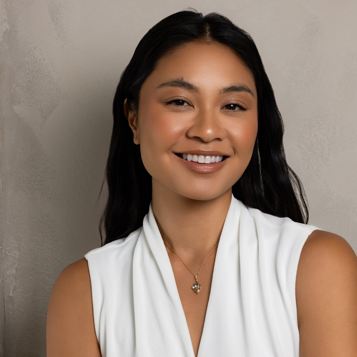 A smiling woman with long dark hair wearing a white sleeveless draped top and a delicate gold necklace, posing with her arms crossed against a textured gray studio background.