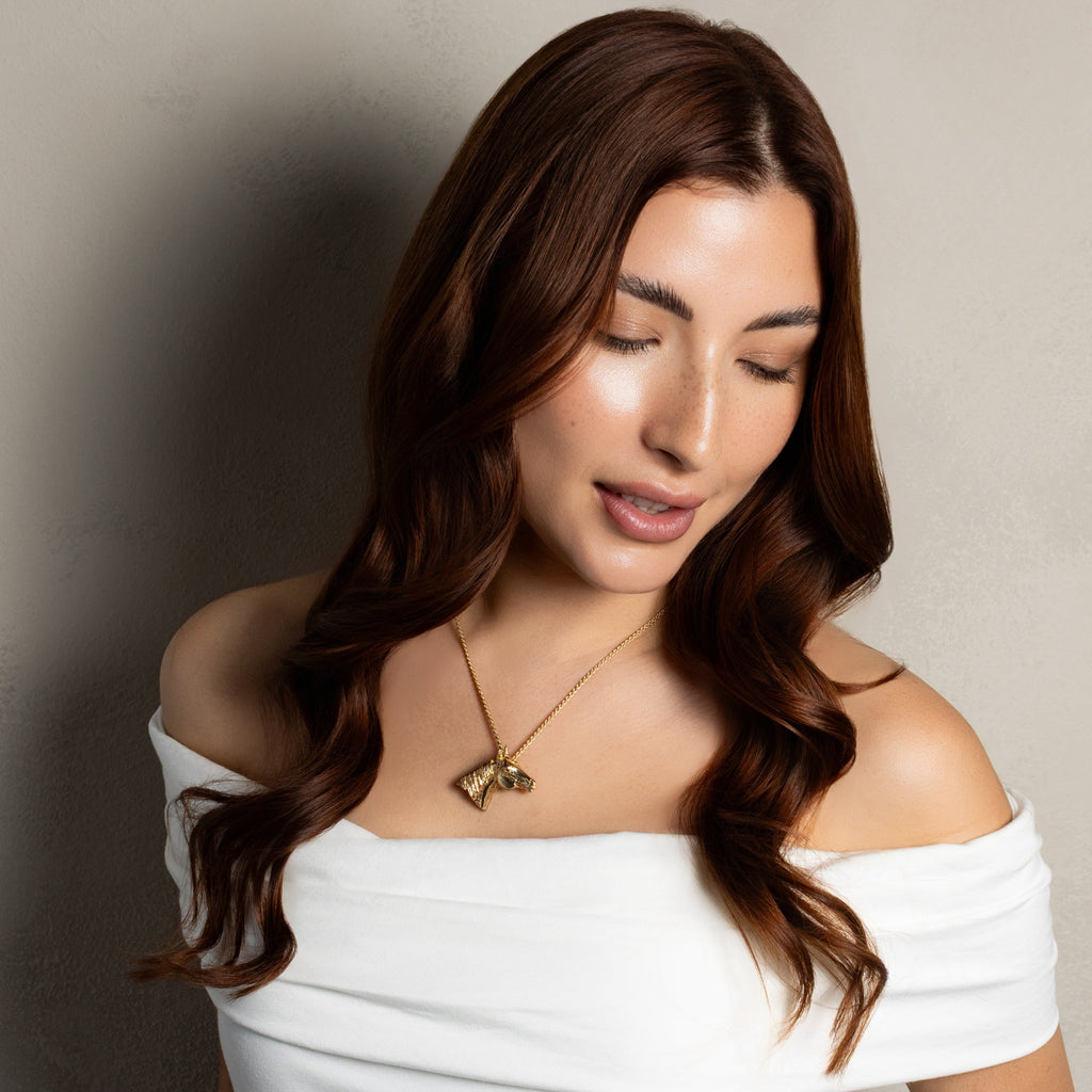 A woman with long, wavy brown hair wearing an off-the-shoulder white top and a gold pendant necklace, posing against a neutral gray textured wall.