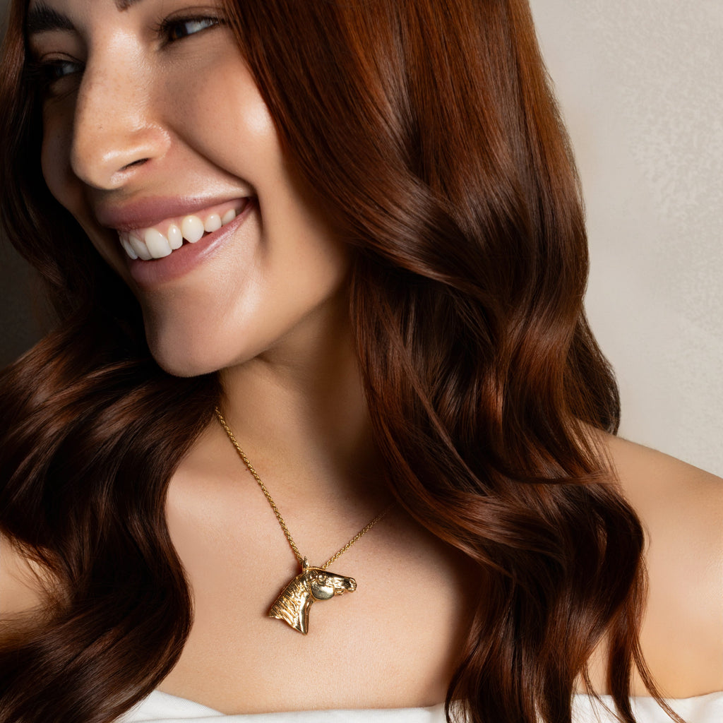 A close-up lifestyle shot of a woman with long, wavy brown hair wearing a gold horse-shaped pendant necklace and a white off-the-shoulder top.