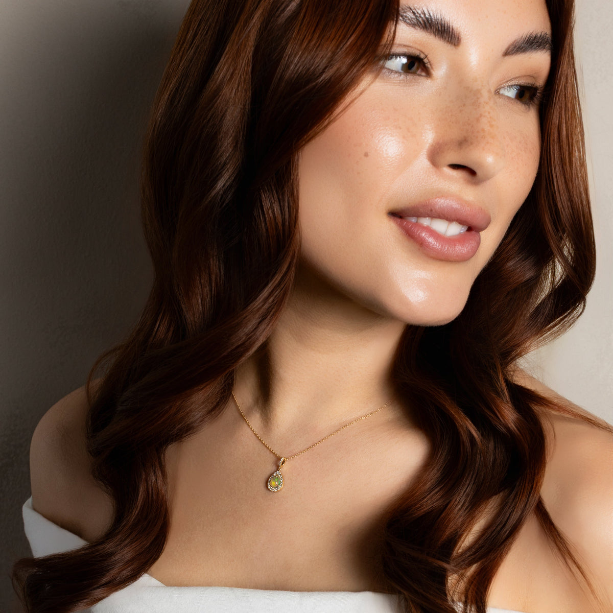 A close-up lifestyle shot of a woman with long, wavy brown hair wearing a delicate gold necklace with a small green stone pendant, paired with a white off-the-shoulder top.