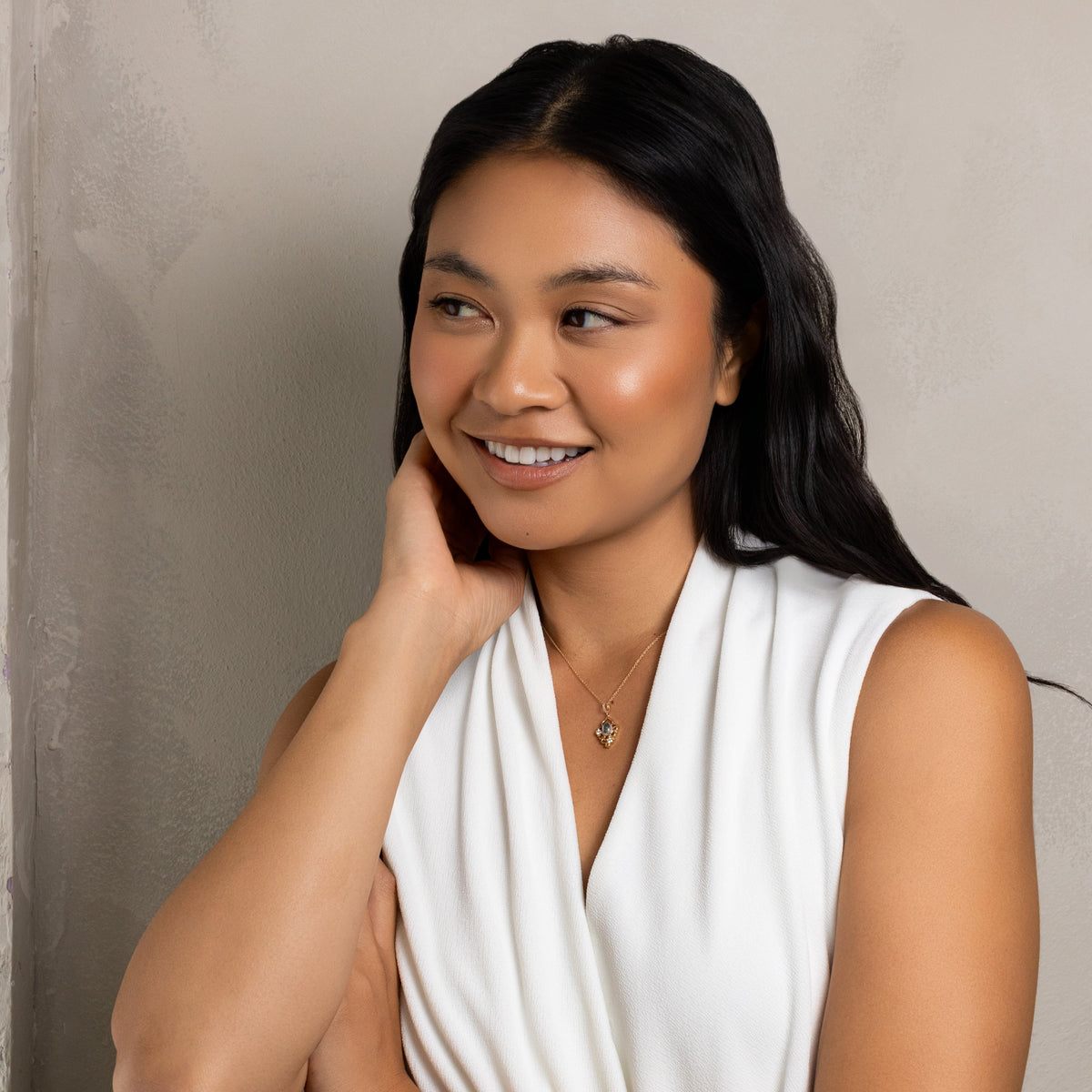 A woman with long dark hair wearing a white sleeveless draped dress, posing against a textured light gray wall.