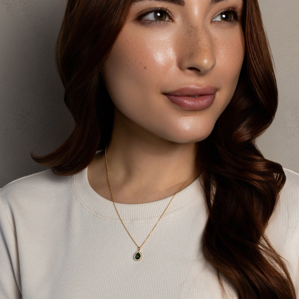 A close-up of a woman wearing a delicate gold necklace with a small dark oval pendant, paired with a ribbed off-white short-sleeve top against a neutral gray background.