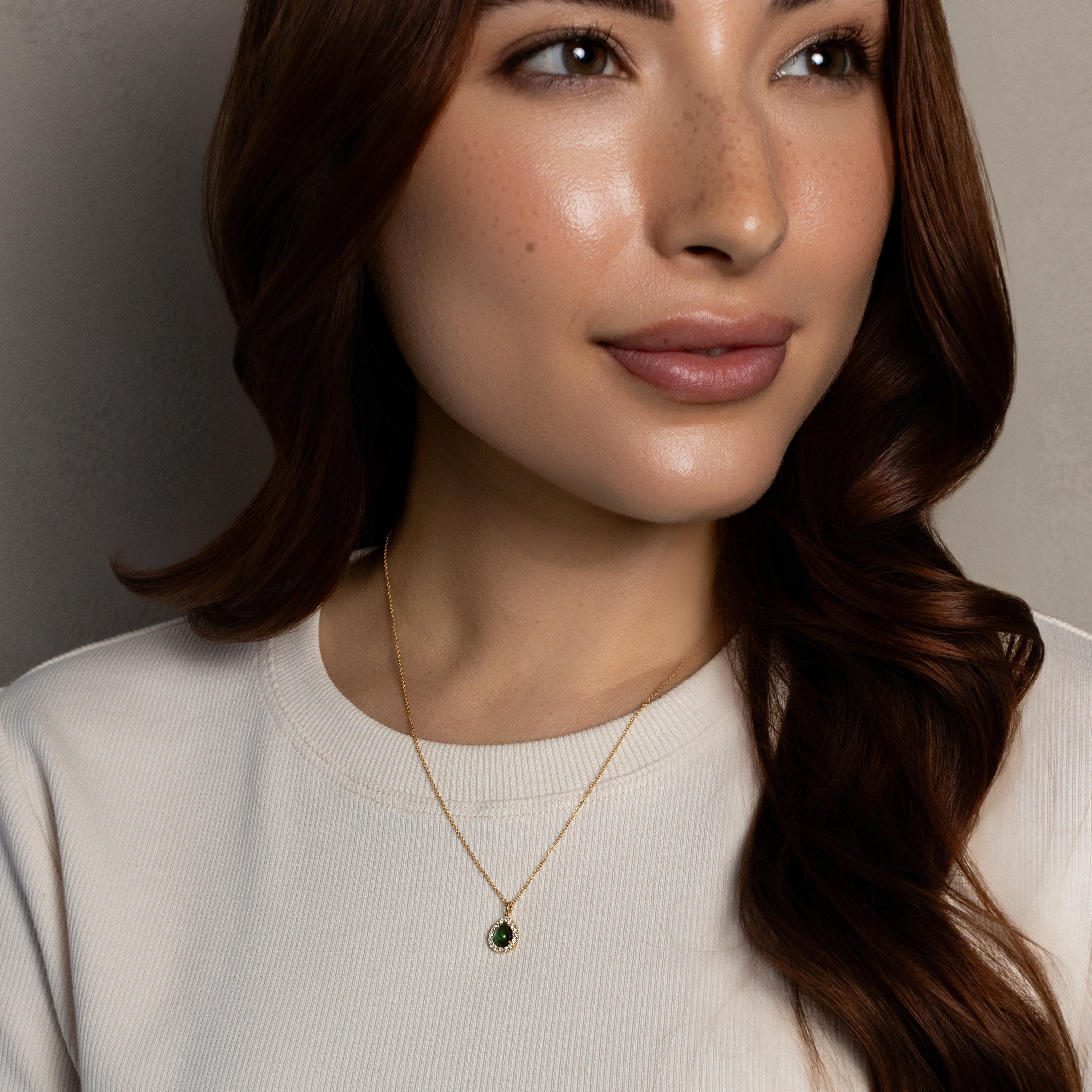 A close-up of a woman wearing a delicate gold necklace with a small dark oval pendant, paired with a ribbed off-white short-sleeve top against a neutral gray background.