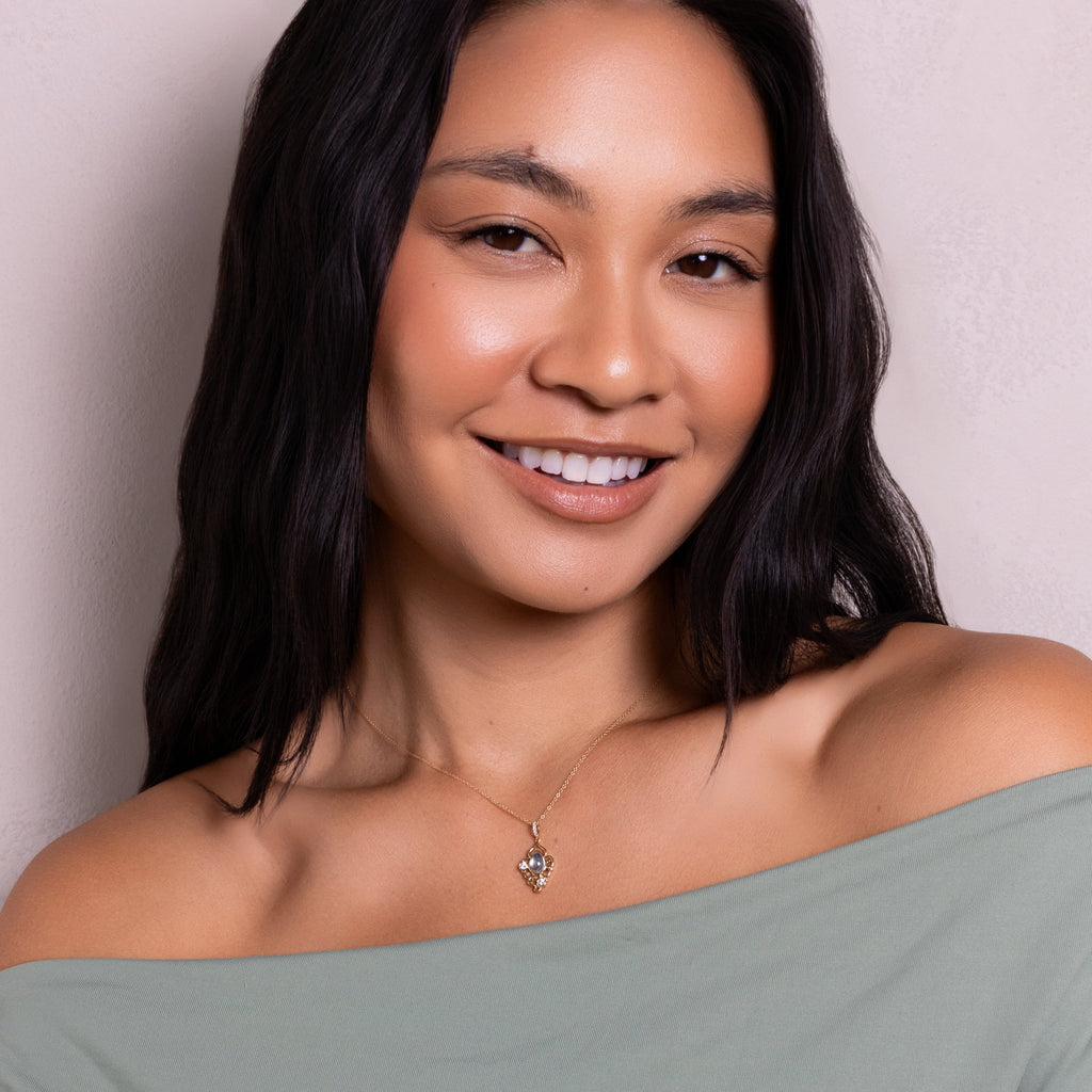 A smiling woman with long dark hair wears an off-the-shoulder sage green top and a delicate heart-shaped pendant necklace, posing against a neutral light beige wall.