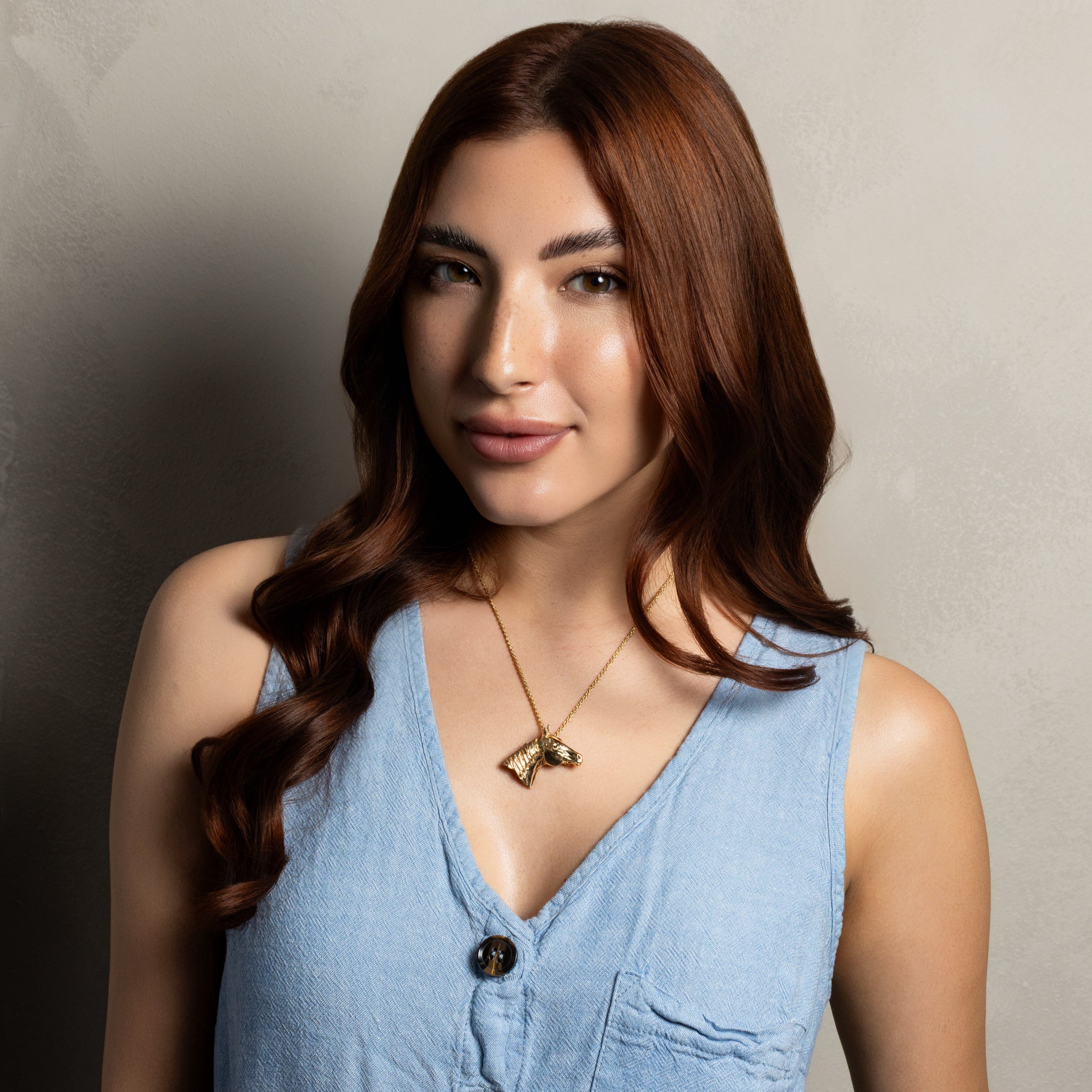 A woman with long brown hair wearing a light blue sleeveless button-front top and a gold pendant necklace, standing against a textured light gray wall.