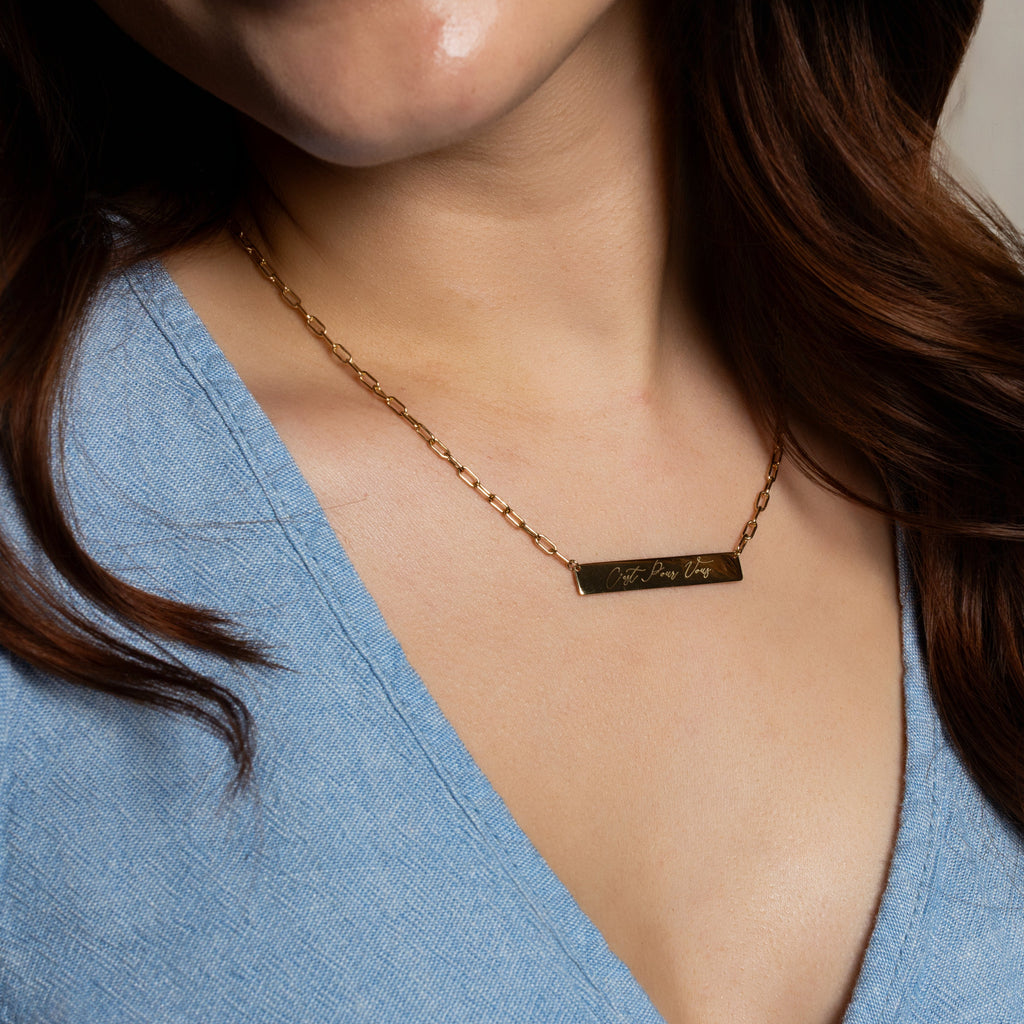 A close-up of a woman wearing a delicate gold chain necklace with a small rectangular bar engraved with the words 'I LOVE YOU' in gold lettering, paired with a light blue V-neck top.