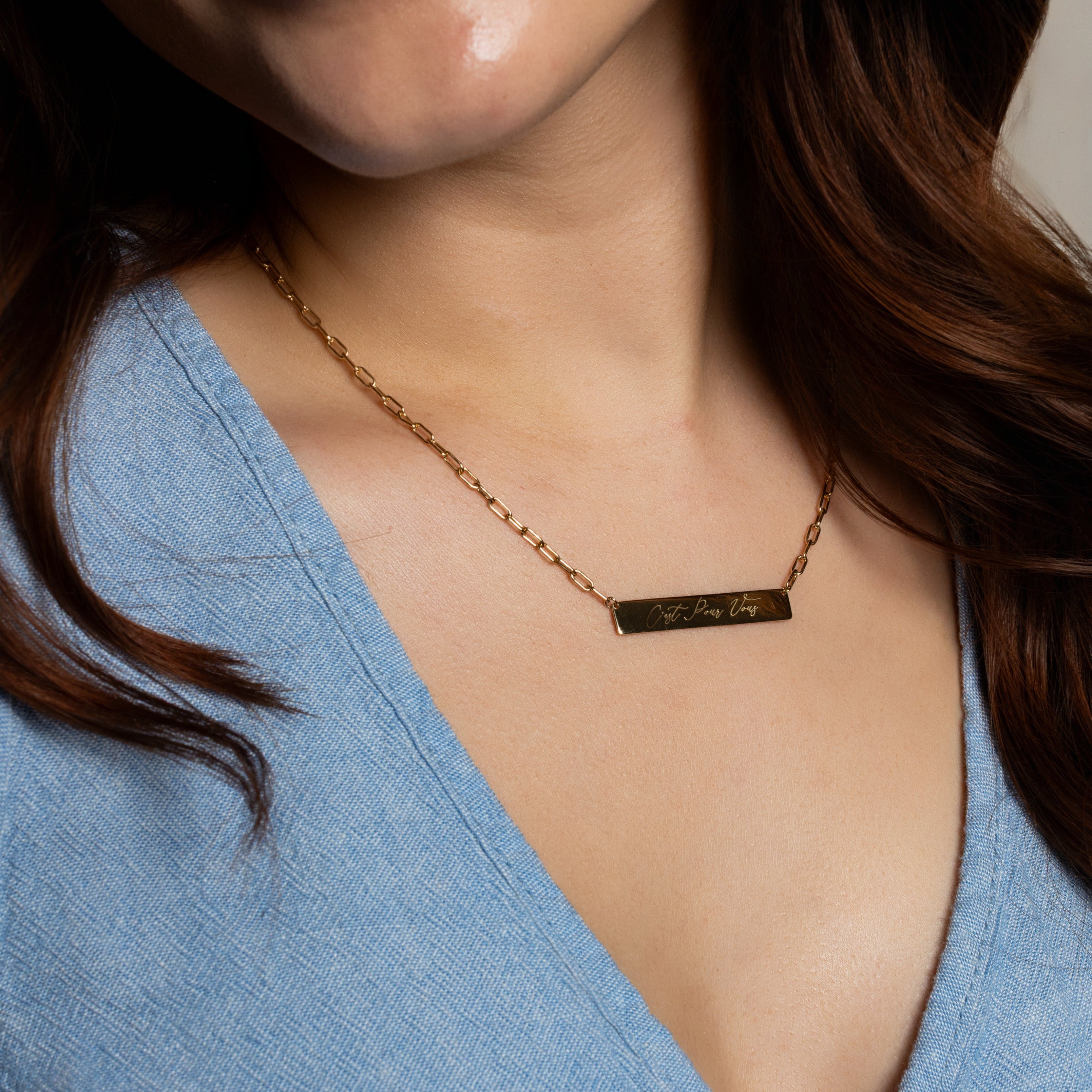 A close-up of a woman wearing a delicate gold chain necklace with a small rectangular bar engraved with the words 'I LOVE YOU' in gold lettering, paired with a light blue V-neck top.