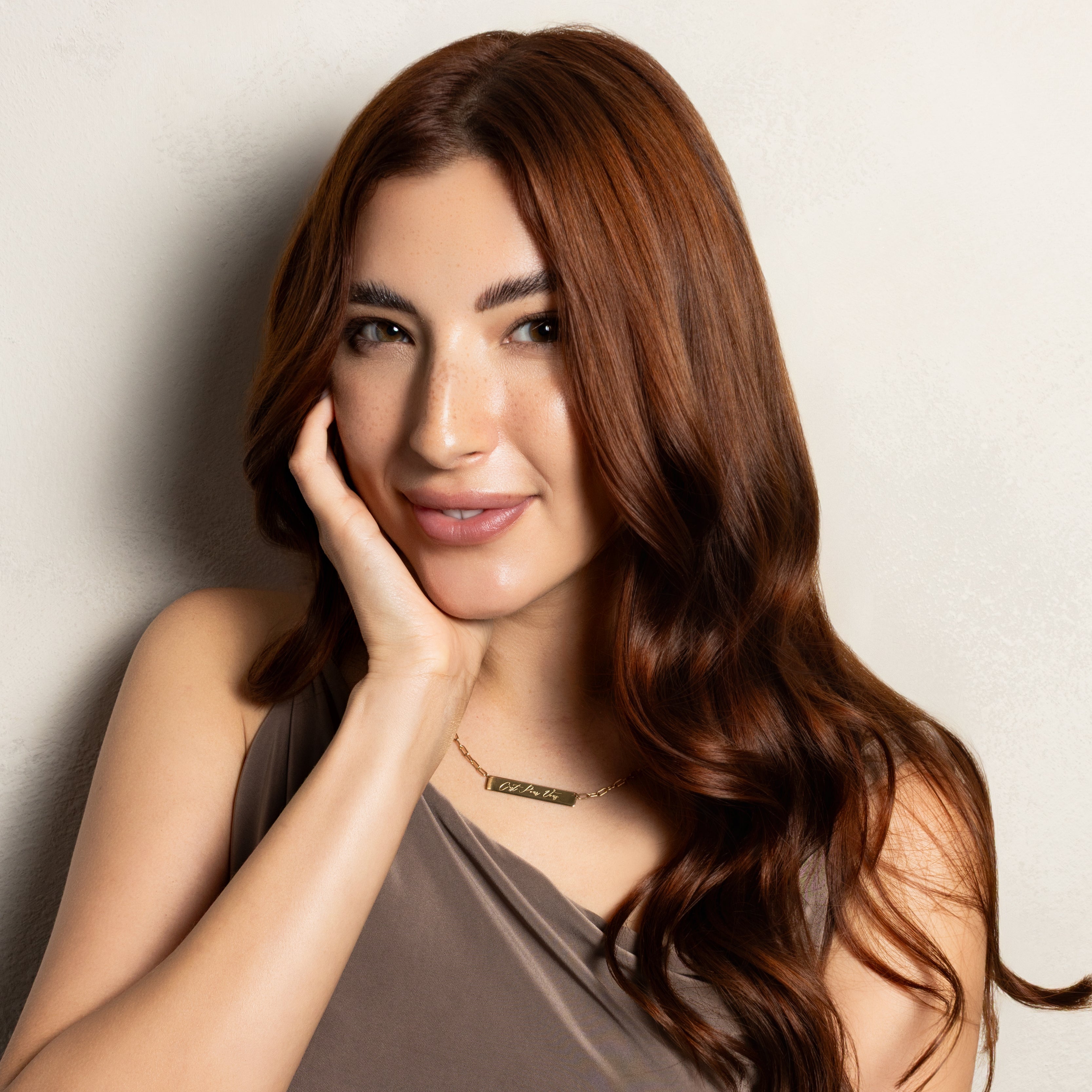 A woman with long, wavy brown hair posing in a taupe-brown one-shoulder top against a light gray textured wall.