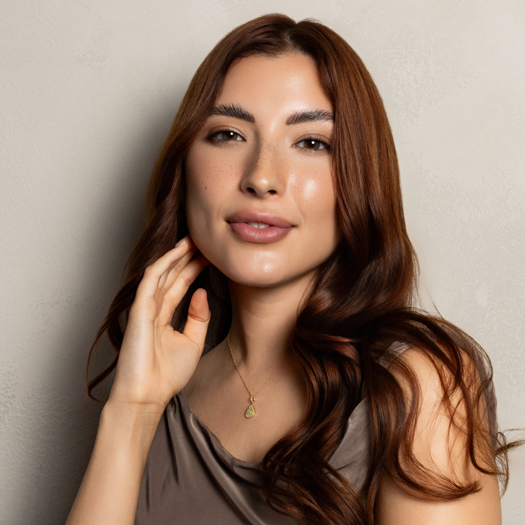 A woman with long, wavy brown hair is shown from the chest up against a plain light gray wall. She is wearing a sleeveless taupe-brown top and a delicate gold necklace with a small green pendant.
